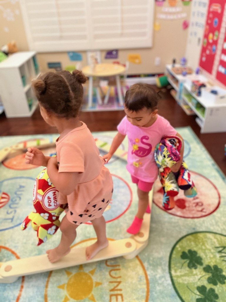 Two toddlers carrying plush toys as they balance along a wooden beam in the PreSchool Prep playroom