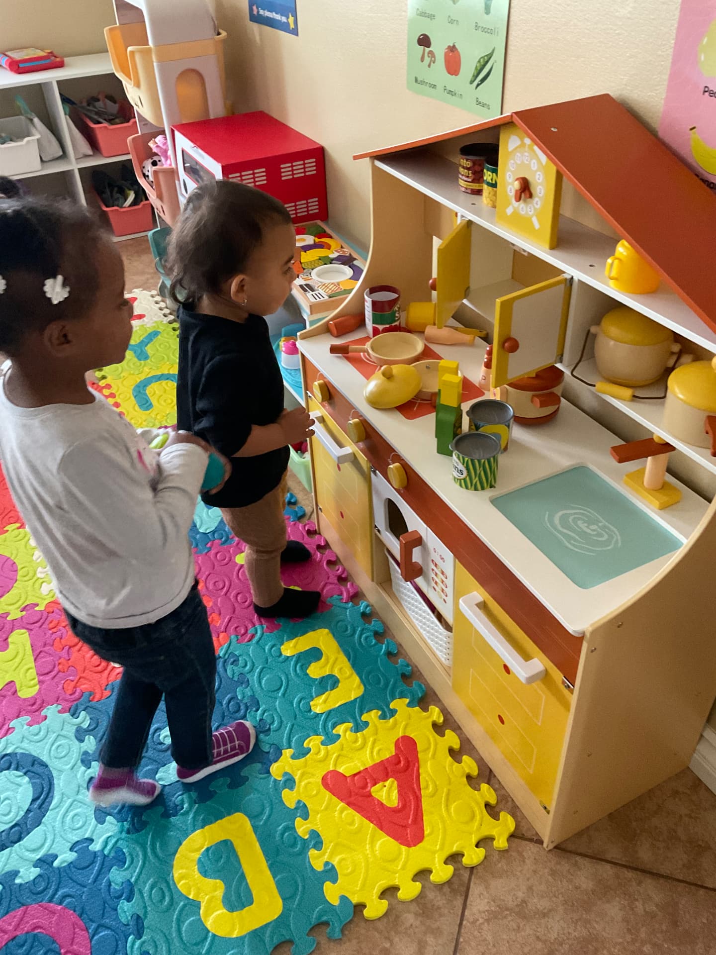Two toddlers exploring the wooden play kitchen on a foam letter mat