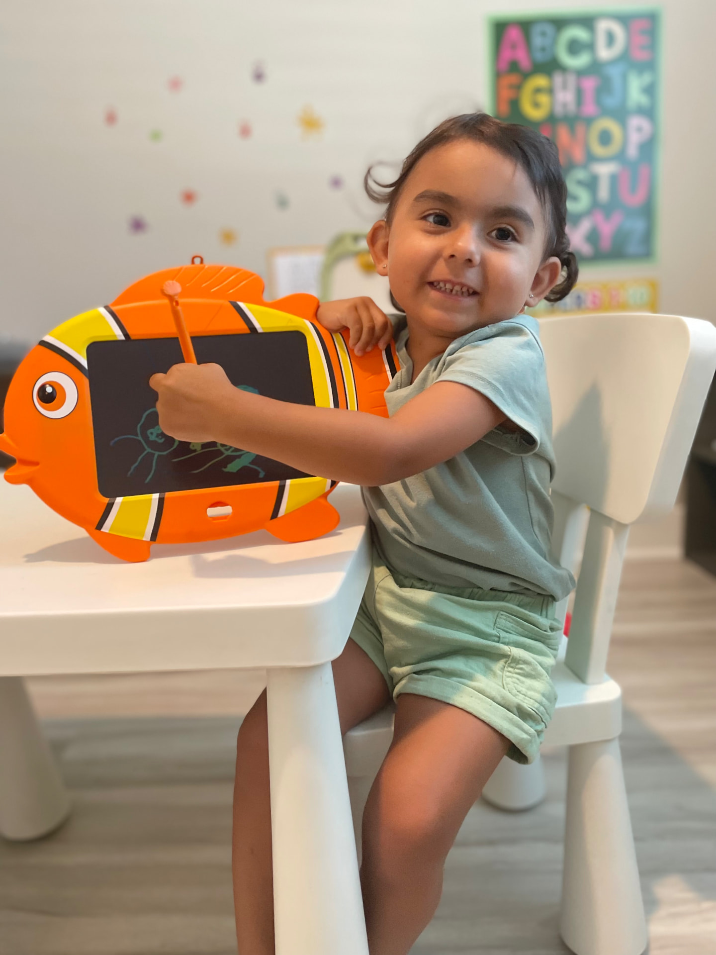 Smiling toddler drawing on a fish-shaped doodle board beside an alphabet poster