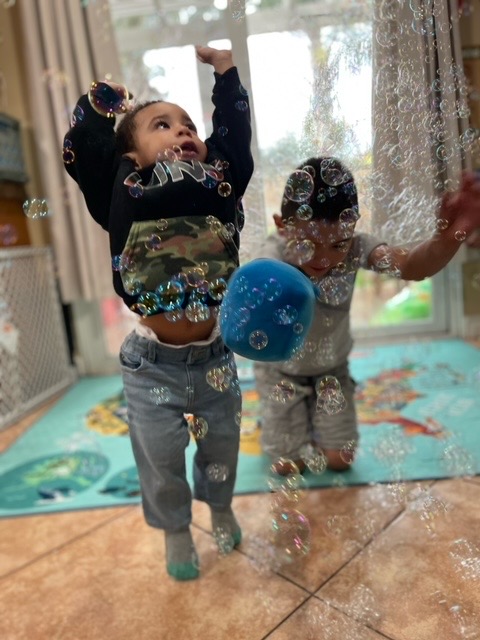 Two toddlers reaching for bubbles floating in the sunlit playroom
