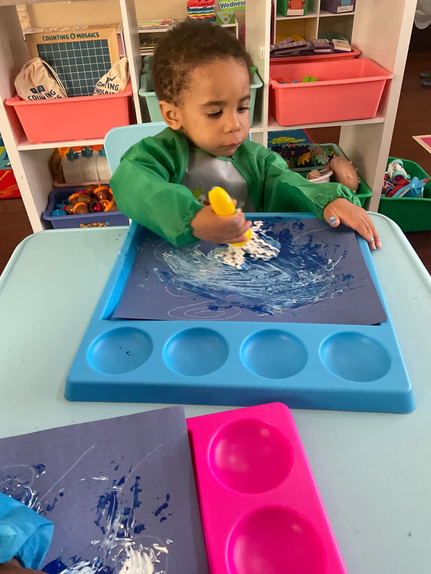 Toddler in a green smock rolling blue paint onto paper at the art table