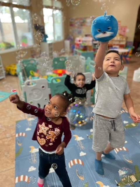 Two toddlers reaching up for bubbles and a plush ball in the playroom